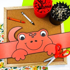 May include: A craft project featuring a red frog headband on a corkboard. Crayons in red and black are in containers. An orange crayon and a white crayon are on the board. Red scissors are on the right. The background is a mix of green and floral patterns.