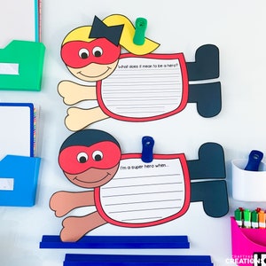 May include: Two superhero character cutouts with blank writing spaces. The top cutout is a girl with blonde hair and a red mask. The bottom cutout is a person with brown skin and a red mask. The text on the cutouts reads "What does it mean to be a hero?" and "I'm a super hero when..."