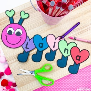 May include: A pink and black caterpillar made of paper with a smiling face and heart-shaped segments. Each heart segment has a letter on it, spelling out the word "Laina".