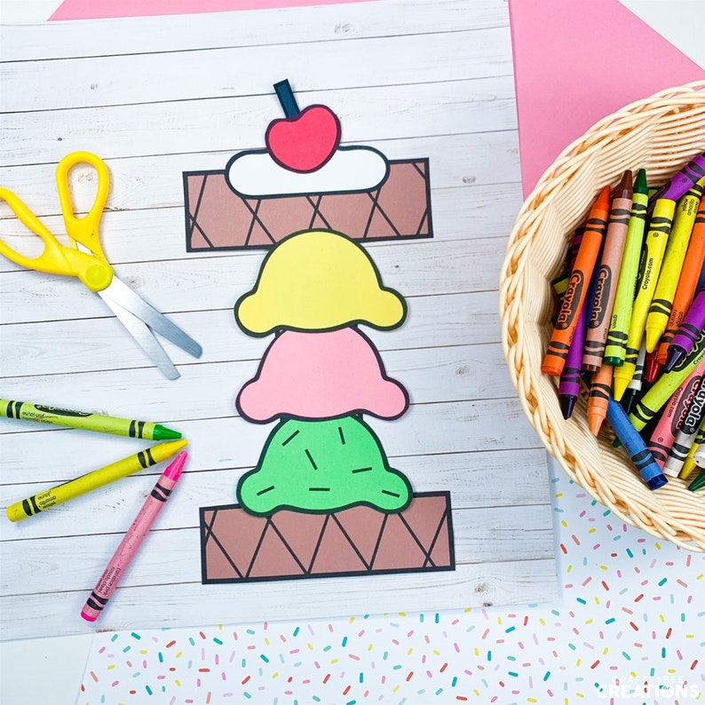 Puede incluir: Un cono de helado de papel artesanal con tres bolas de helado: amarillo, rosa y verde. El cono de helado est&aacute; sobre un fondo de madera blanca con una cesta de l&aacute;pices de colores en el fondo.