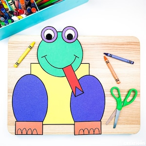 May include: A paper craft frog made with different colored paper shapes. The frog is green with a red tongue, yellow body, and purple legs. The frog is sitting on a wooden surface with crayons and scissors.