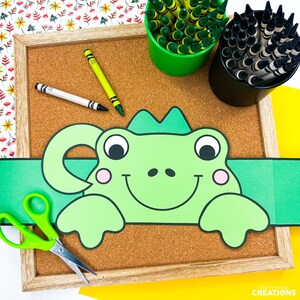 Puede incluir: Una manualidad de diadema con forma de rana verde sobre un corcho, con tijeras verdes, crayones y un crayón blanco y amarillo. La diadema tiene una cara sonriente y mejillas rosadas. El fondo incluye un estampado floral y papel amarillo.