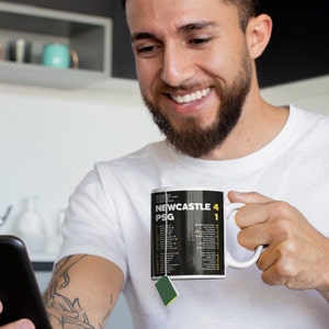 K&ouml;nnte beinhalten: Wei&szlig;e Keramik-Tasse mit dem Text "NEWCASTLE 4 PSG 1" in Gelb und Wei&szlig; auf schwarzem Hintergrund. Die Tasse wird von einer Person gehalten, die ein wei&szlig;es T-Shirt tr&auml;gt.