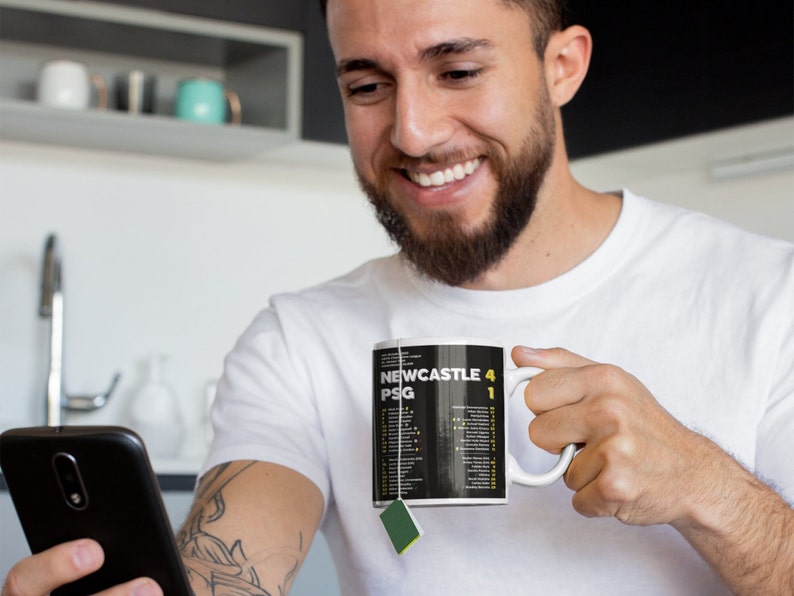 K&ouml;nnte beinhalten: Wei&szlig;e Keramik-Tasse mit dem Text "NEWCASTLE 4 PSG 1" in Gelb und Wei&szlig; auf schwarzem Hintergrund. Die Tasse wird von einer Person gehalten, die ein wei&szlig;es T-Shirt tr&auml;gt.