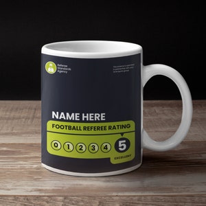 May include: White ceramic mug with a black and green design. The mug features the text "NAME HERE FOOTBALL REFEREE RATING" and a rating scale from 0 to 5, with 5 being "EXCELLENT".