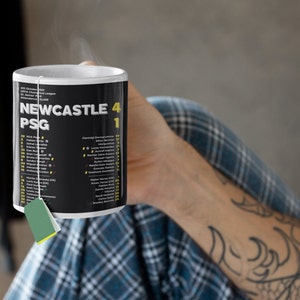 K&ouml;nnte beinhalten: Schwarz-wei&szlig;e Keramik-Tasse mit der Aufschrift "NEWCASTLE 4 PSG 1" und Spielernamen. Die Tasse wird von einer Person gehalten und hat einen Teebeutel. Ein Sportartikel.