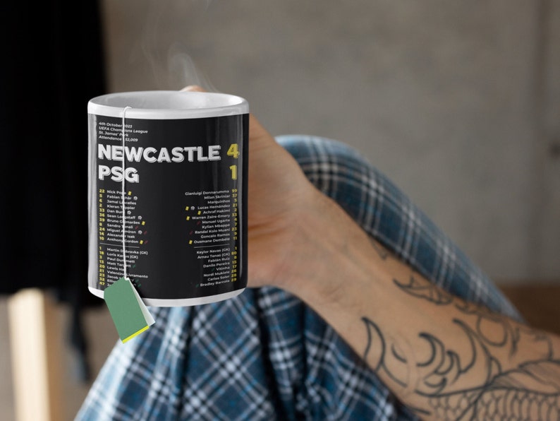 K&ouml;nnte beinhalten: Schwarz-wei&szlig;e Keramik-Tasse mit der Aufschrift "NEWCASTLE 4 PSG 1" und Spielernamen. Die Tasse wird von einer Person gehalten und hat einen Teebeutel. Ein Sportartikel.