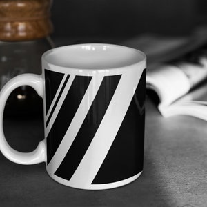 Puede incluir: Una taza de cer&aacute;mica blanca con un dise&ntilde;o de rayas blancas y negras. La taza tiene un asa y est&aacute; sobre una superficie gris.