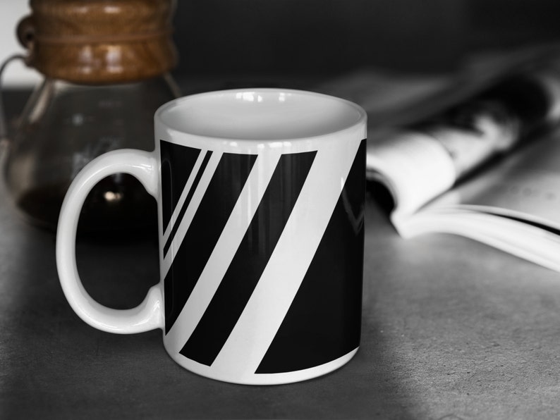 Puede incluir: Una taza de cer&aacute;mica blanca con un dise&ntilde;o de rayas blancas y negras. La taza tiene un asa y est&aacute; sobre una superficie gris.
