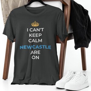 May include: A dark grey t-shirt with a white crown graphic and the text "I can't keep calm Newcastle are on".
