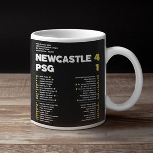 K&ouml;nnte beinhalten: Eine wei&szlig;e Keramik-Tasse mit schwarzem Design, die den Text "NEWCASTLE 4 PSG 1" zeigt. Die Tasse enth&auml;lt Spielernamen und Spielinformationen. Sie hat einen wei&szlig;en Henkel und steht vor einem dunklen Hintergrund, f&uuml;r Fu&szlig;ballfans.