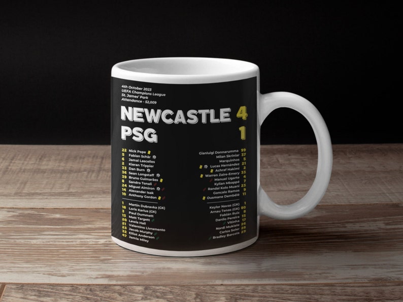 K&ouml;nnte beinhalten: Eine wei&szlig;e Keramik-Tasse mit schwarzem Design, die den Text "NEWCASTLE 4 PSG 1" zeigt. Die Tasse enth&auml;lt Spielernamen und Spielinformationen. Sie hat einen wei&szlig;en Henkel und steht vor einem dunklen Hintergrund, f&uuml;r Fu&szlig;ballfans.