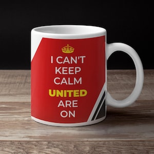 Può includere: Tazza in ceramica bianca con un design rosso, nero e bianco. La tazza ha il testo "I can't keep calm United are on" in lettere gialle e nere.