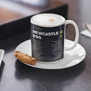 K&ouml;nnte beinhalten: Eine wei&szlig;e Keramiktasse mit einem schwarzen Band mit der Aufschrift "NEWCASTLE 4 PSG 1" und Spielernamen. Die Tasse steht auf einer wei&szlig;en Untertasse, daneben Kekse. Die Tasse enth&auml;lt ein schaumiges Getr&auml;nk.