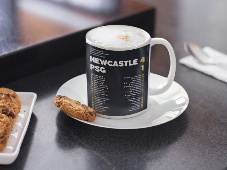 K&ouml;nnte beinhalten: Eine wei&szlig;e Keramiktasse mit einem schwarzen Band mit der Aufschrift "NEWCASTLE 4 PSG 1" und Spielernamen. Die Tasse steht auf einer wei&szlig;en Untertasse, daneben Kekse. Die Tasse enth&auml;lt ein schaumiges Getr&auml;nk.