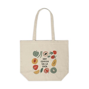 May include: A beige tote bag with a colorful illustration of fruits and vegetables. The text "Hay comida en la casa" is printed on the bag.