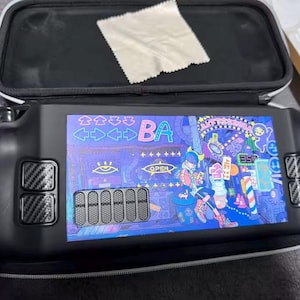 Puede incluir: Dispositivo de juego portátil negro con una pantalla vibrante que muestra una colorida ilustración de estilo anime. El dispositivo está en una funda negra con cremallera blanca. La pantalla muestra la palabra "BA" y otros textos. Un paño de limpieza descansa sobre la funda.