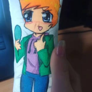 May include: A small, rectangular pillow with a cartoon illustration of a character with orange hair, blue eyes, and a green jacket. The character is holding a teal object. The pillow has a white background. The character is wearing a purple hoodie.
