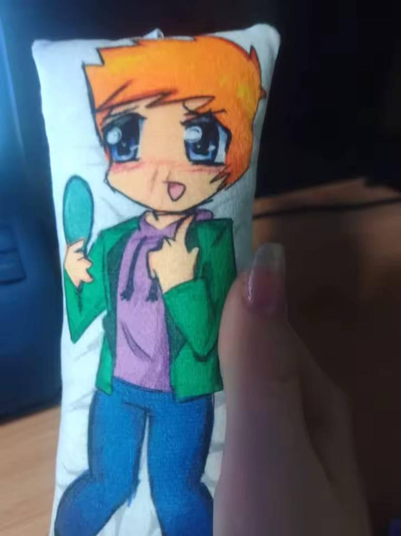 May include: A small, rectangular pillow with a cartoon illustration of a character with orange hair, blue eyes, and a green jacket. The character is holding a teal object. The pillow has a white background. The character is wearing a purple hoodie.