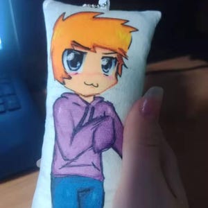 May include: A rectangular plush keychain with a cartoon character design. The character has orange hair, a purple hoodie, and blue jeans. The character has large eyes and a small smile. A silver metal loop is attached.
