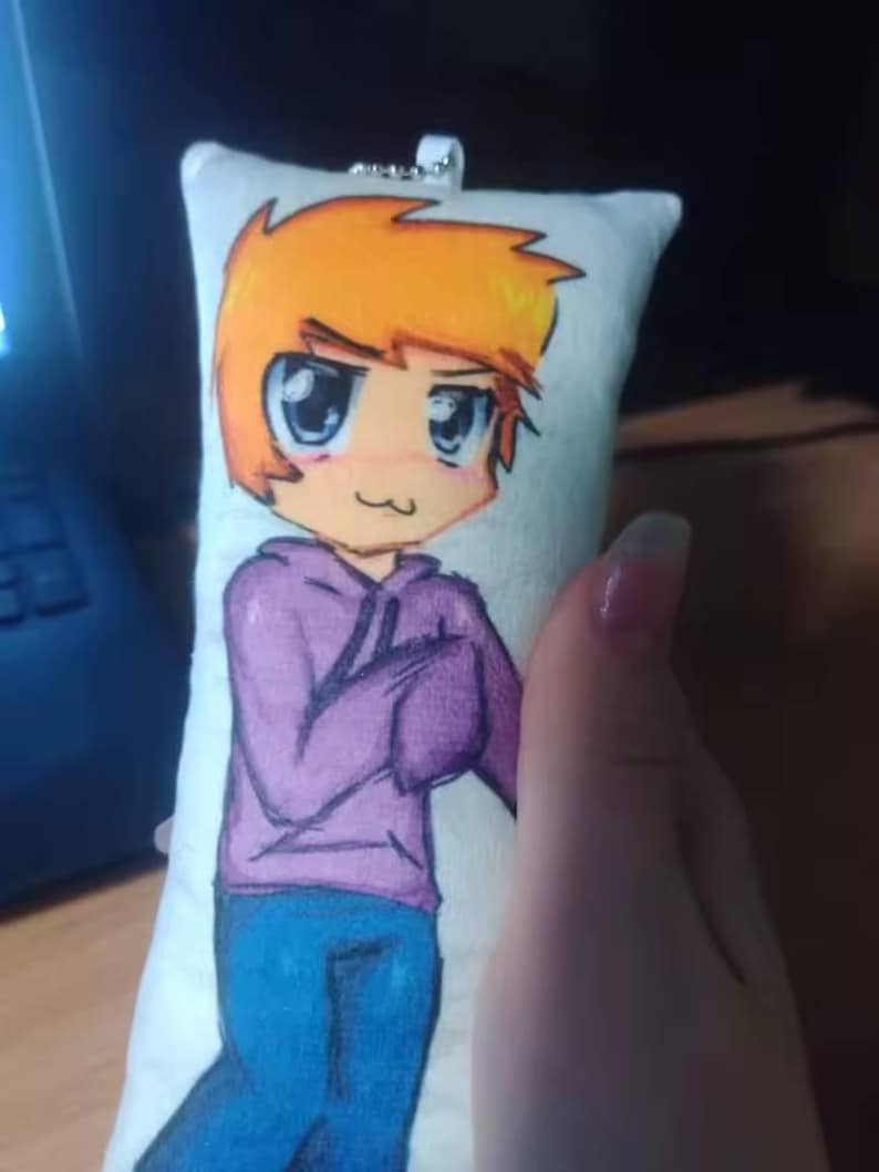 May include: A rectangular plush keychain with a cartoon character design. The character has orange hair, a purple hoodie, and blue jeans. The character has large eyes and a small smile. A silver metal loop is attached.