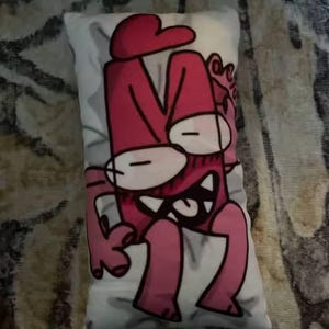 May include: A rectangular cushion with a cartoon character design in pink and white. The character has a large head with a hat, wide eyes, and a toothy grin. The cushion is placed on a patterned surface.