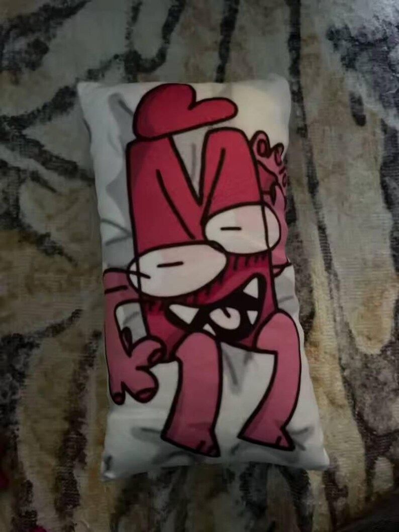 May include: A rectangular cushion with a cartoon character design in pink and white. The character has a large head with a hat, wide eyes, and a toothy grin. The cushion is placed on a patterned surface.