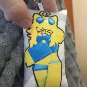 May include: A small, rectangular cushion featuring a cartoon character. The character is yellow with blue details, wearing sunglasses, and smiling. The cushion is attached to a silver chain.