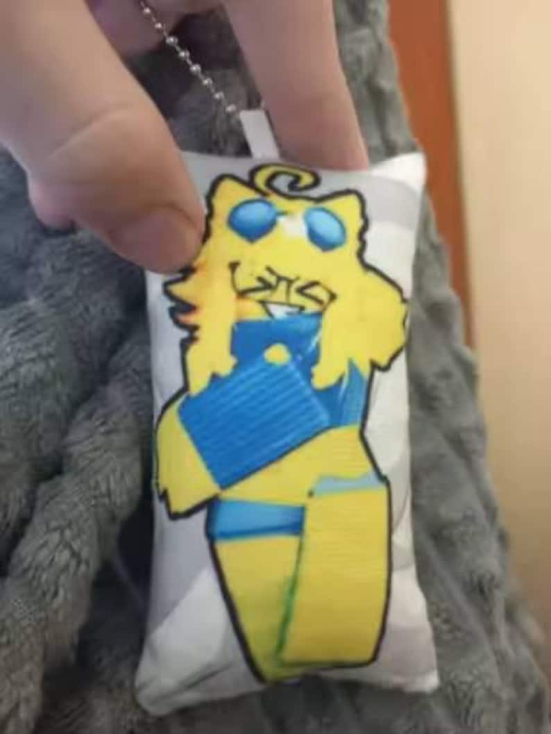 May include: A small, rectangular cushion featuring a cartoon character. The character is yellow with blue details, wearing sunglasses, and smiling. The cushion is attached to a silver chain.