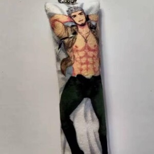 May include: A rectangular pillow-shaped keyring with a printed image of a muscular person in a tan jacket, black trousers, and black boots. The person is reclining with arms behind their head. The keyring has a silver metal clasp.