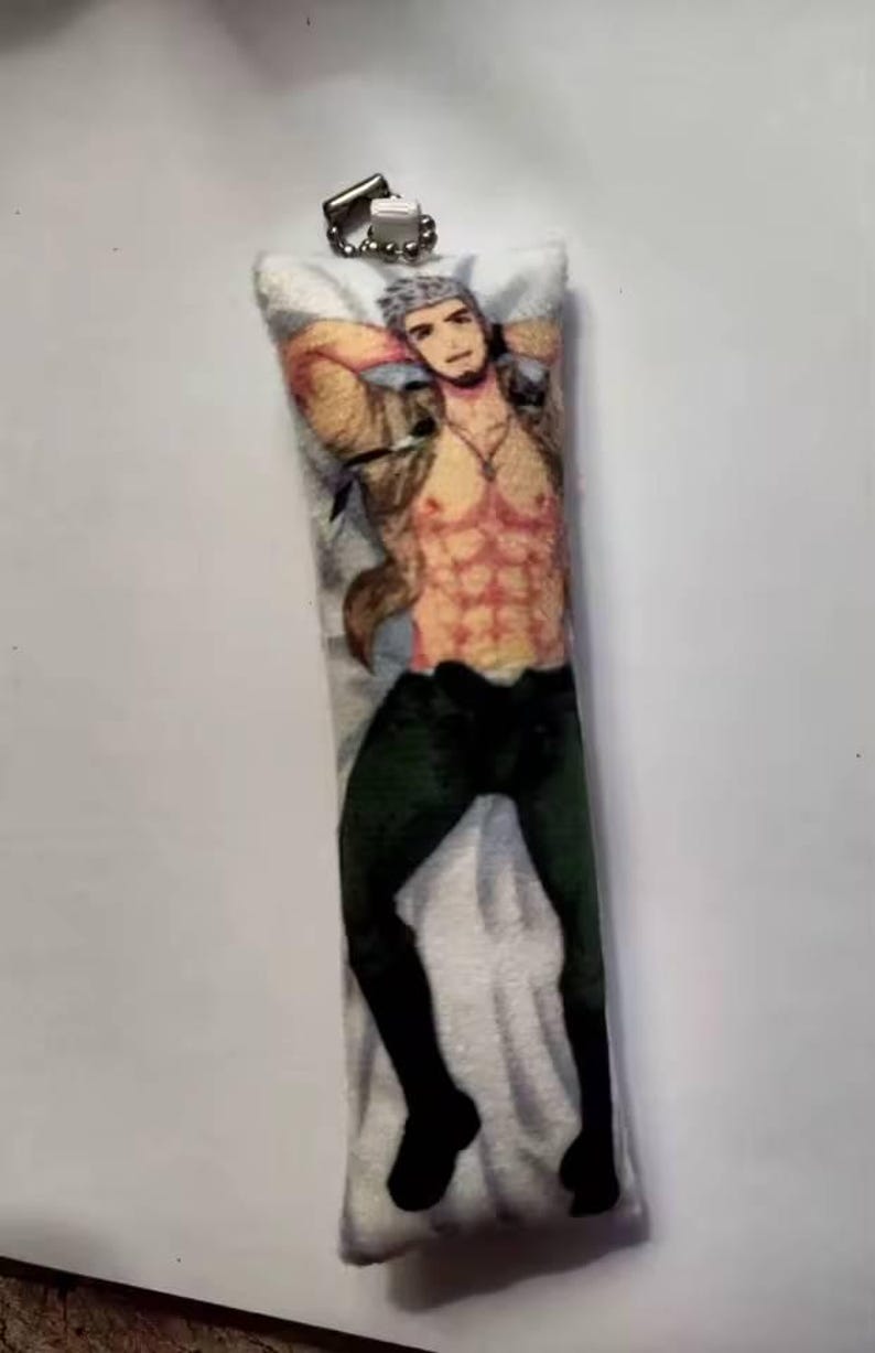 May include: A rectangular pillow-shaped keyring with a printed image of a muscular person in a tan jacket, black trousers, and black boots. The person is reclining with arms behind their head. The keyring has a silver metal clasp.
