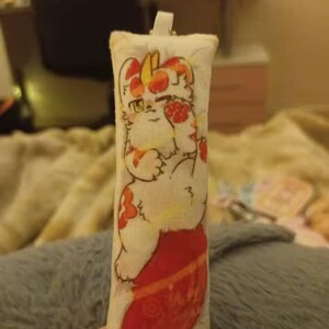 May include: A rectangular, cream-coloured plush keychain with a cartoon cat design in red and orange. The cat is shown with a red stocking. The keychain has a white loop at the top. The date and time 2025/12/26 21:37 and the symbol <3 are printed at the bottom.