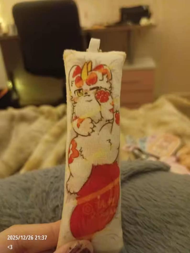 May include: A rectangular, cream-coloured plush keychain with a cartoon cat design in red and orange. The cat is shown with a red stocking. The keychain has a white loop at the top. The date and time 2025/12/26 21:37 and the symbol <3 are printed at the bottom.