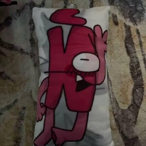 May include: A rectangular white cushion with a cartoon character in red and pink. The character has a large red head, a single white eye, and red shorts. A silver chain is attached to the top of the cushion.