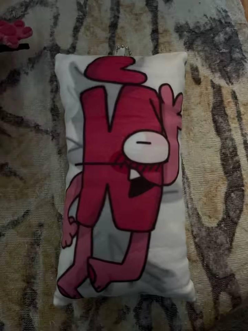 May include: A rectangular white cushion with a cartoon character in red and pink. The character has a large red head, a single white eye, and red shorts. A silver chain is attached to the top of the cushion.
