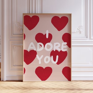 May include: Framed art print with a light pink background and a pattern of red hearts. The words "I ADORE YOU" are written in white. The artwork is displayed in a gold frame.