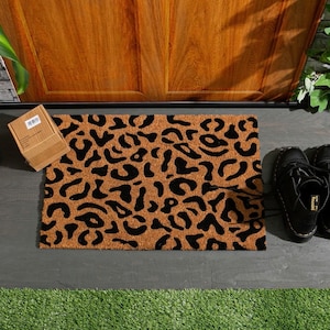 May include: A brown coir doormat with a black leopard print design. The doormat is placed on a gray concrete surface.
