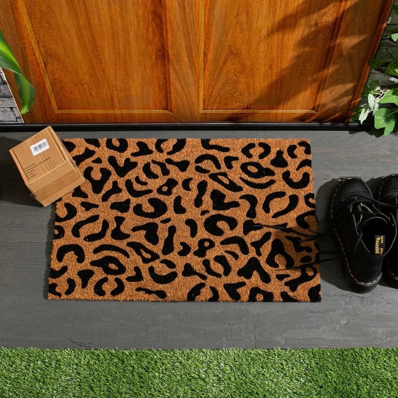 May include: A brown coir doormat with a black leopard print design. The doormat is placed on a gray concrete surface.