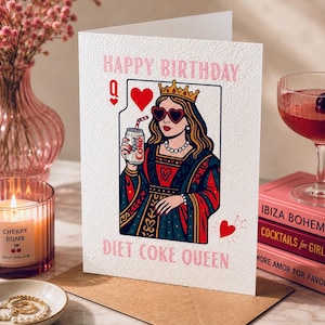 May include: A greeting card with the text "HAPPY BIRTHDAY" and "DIET COKE QUEEN". The card features a queen illustration holding a Diet Coke can, wearing heart-shaped sunglasses and a crown. A lit candle and a glass of pink cocktail are in the background.