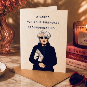 Puede incluir: Una tarjeta de felicitaci&oacute;n plegada con el texto "A CARD? FOR YOUR BIRTHDAY? GROUNDBREAKING..." y una ilustraci&oacute;n de una mujer con gafas de sol sosteniendo una revista. Tambi&eacute;n se ven una vela encendida, libros y gafas de sol en forma de coraz&oacute;n.