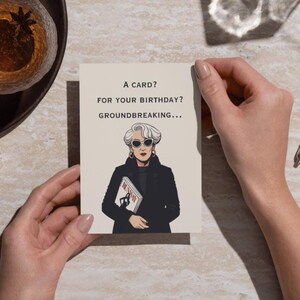 Puede incluir: Tarjeta de felicitaci&oacute;n de cumplea&ntilde;os sostenida por manos, con una ilustraci&oacute;n de una mujer con gafas de sol y abrigo negro. La tarjeta dice: "&iquest;UNA TARJETA? &iquest;PARA TU CUMPLEA&Ntilde;OS? REVOLUCIONARIO..."