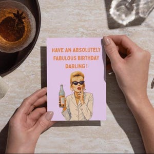 May include: A birthday card with the text "HAVE AN ABSOLUTELY FABULOUS BIRTHDAY DARLING!" features a pop art illustration of a woman in sunglasses holding a bottle. The card is held by a person.