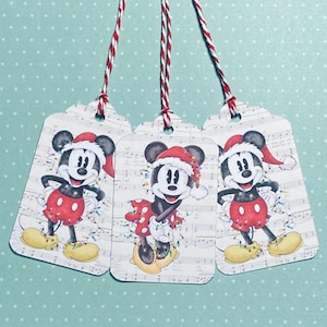 May include: Set of three Christmas gift tags featuring Mickey Mouse wearing a Santa hat and surrounded by Christmas lights. The tags are printed on a white background with musical notes.