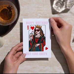 May include: A white greeting card with the text "HAPPY BIRTHDAY" and "DIET COKE QUEEN". The card features a queen illustration wearing heart-shaped sunglasses and holding a Diet Coke can. The card has a playing card design with hearts.