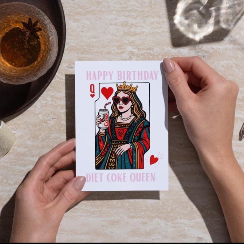 May include: A white greeting card with the text "HAPPY BIRTHDAY" and "DIET COKE QUEEN". The card features a queen illustration wearing heart-shaped sunglasses and holding a Diet Coke can. The card has a playing card design with hearts.
