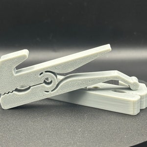Może przedstawiać: Szara plastikowa klamra do bielizny o unikalnym designie. Klamra do bielizny jest wykonana z teksturowanego plastiku i posiada sprężynowy mechanizm.