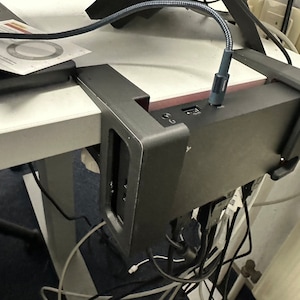Könnte beinhalten: Ein schwarzes Gerät mit mehreren Anschlüssen und angeschlossenen Kabeln, das mit einer Metallhalterung an der Unterseite eines Schreibtisches befestigt ist. Das Gerät ist an eine Steckdose mit der Aufschrift "PC-NETZ" angeschlossen.
