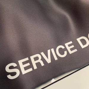 May include: A black fabric item featuring the white text SERVICE D printed in a bold, sans-serif font. The material has a smooth, slightly reflective finish with visible folds and a straight stitched edge along the bottom border of the piece.