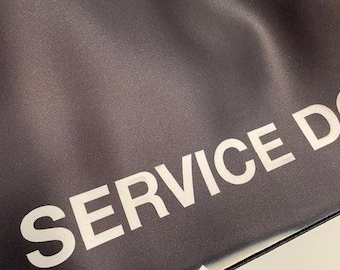 May include: A black fabric item featuring the white text SERVICE D printed in a bold, sans-serif font. The material has a smooth, slightly reflective finish with visible folds and a straight stitched edge along the bottom border of the piece.