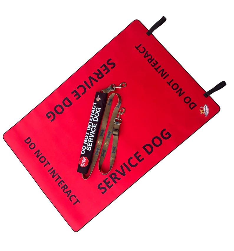 May include: A rectangular red mat with black text reading SERVICE DOG and DO NOT INTERACT, featuring black corner tabs. Included is a black leash wrap with the text DO NOT INTERACT SERVICE DOG and a stop sign icon, plus a tan leash with bone patterns.